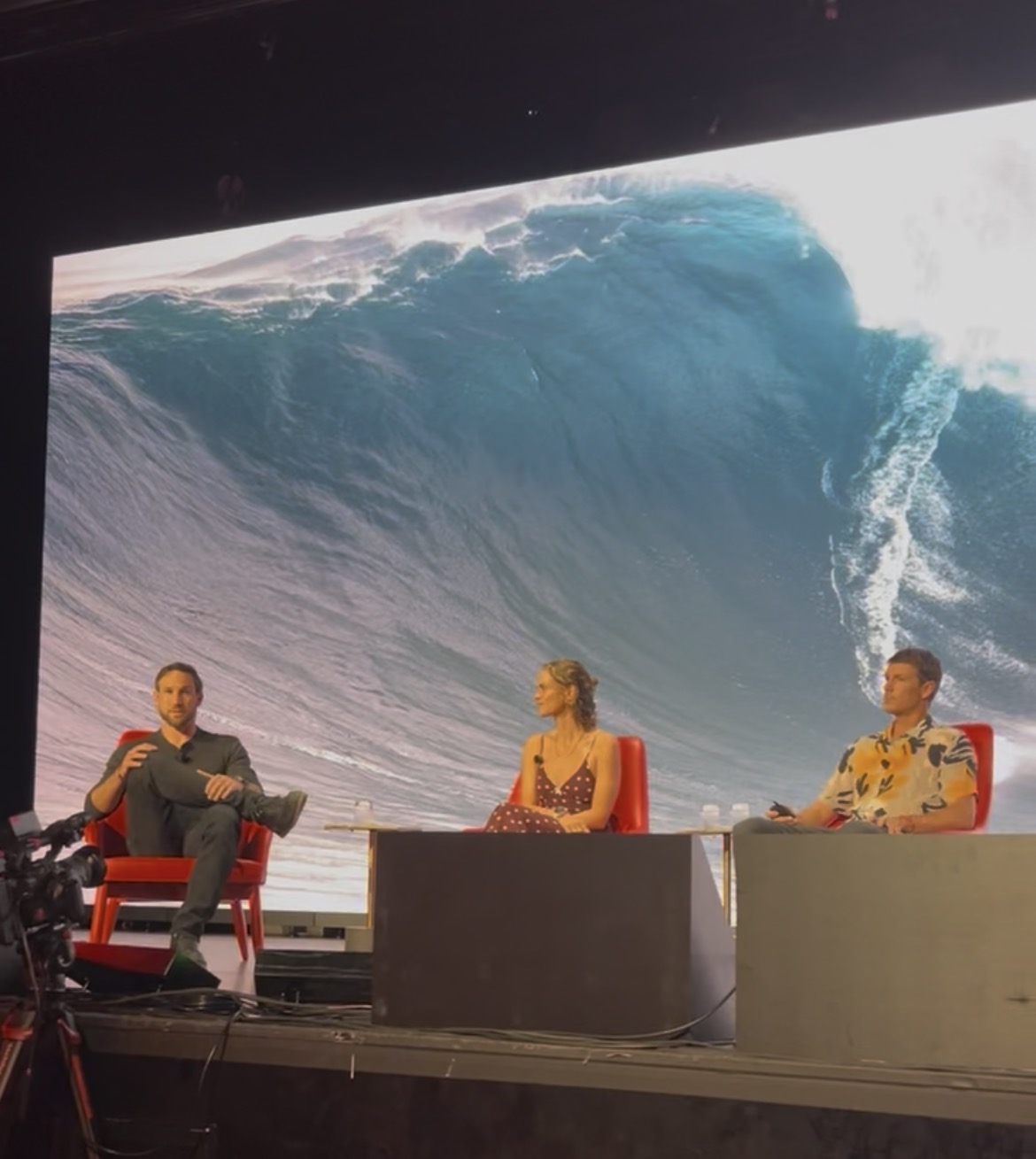 Panel discussion on stage, ocean wave projected behind